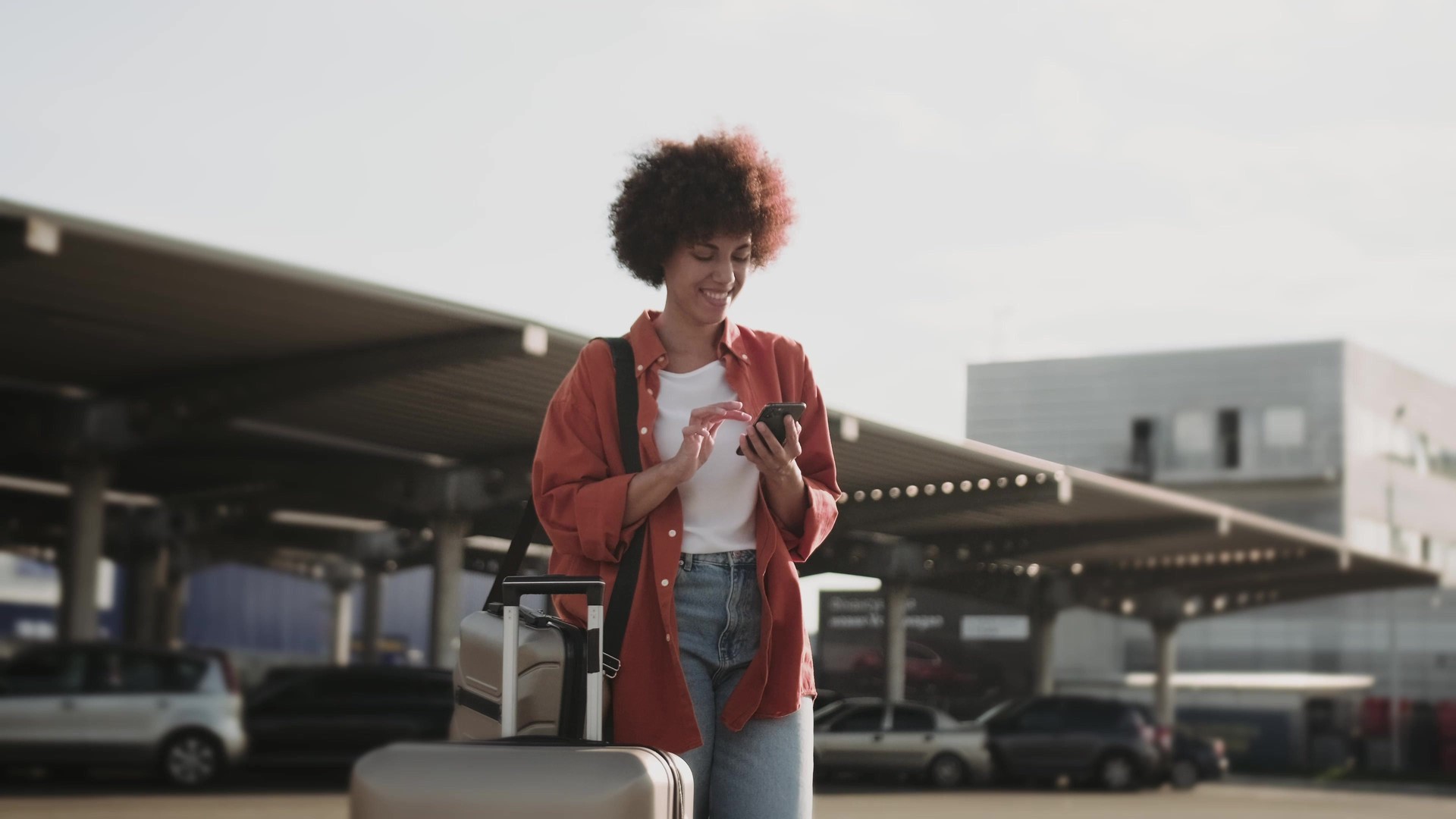 Happy female with luggage typing on the mobile phone outside.